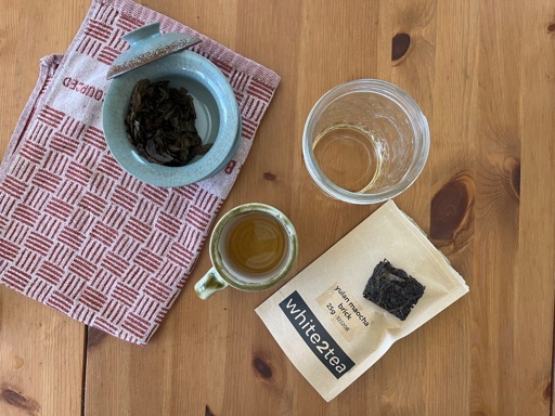 Tea in a gaiwan, with a cup and serving vessel filled with tea, and a bag of tea labeled “yulan maocha brick, 25g, White2Tea”