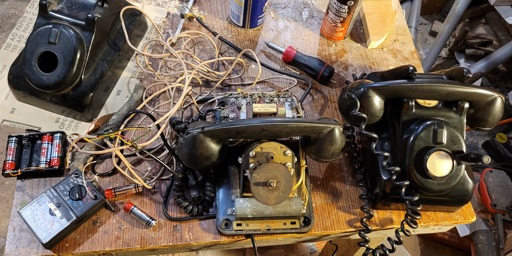 A cluttered workshop with two antique phones, one partially disassembled, linked with a tangle of wire and some janky connections to a battery pack