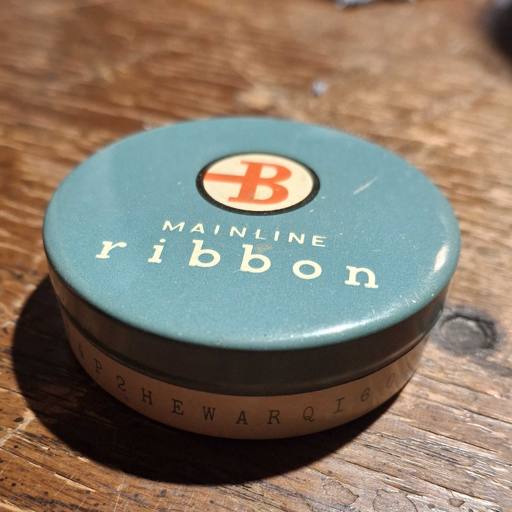 A small, round metal tin. The lid is blue with the title "MAINLINE ribbon" and a company monogram of "B". The body is beige with blue text wrapped around the container.